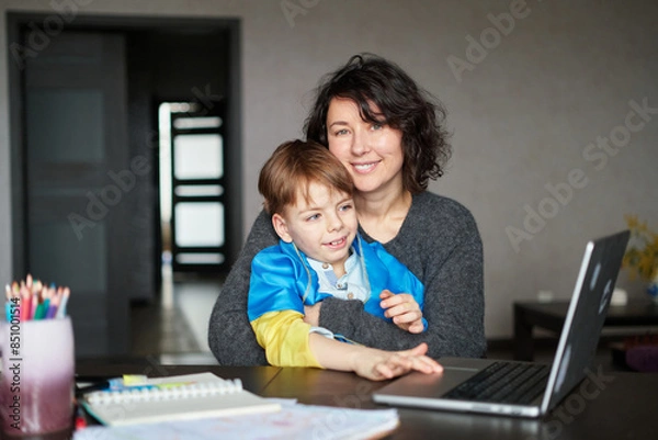 Obraz Ukrainian mother and son with flag sitting with laptop at home studying .Happy smiling family hugs and looking at the camera. Ukrainian refugees concept. High quality photo