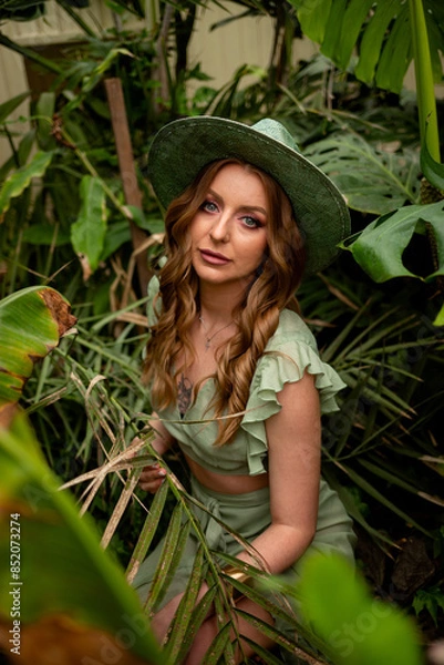 Obraz young brunette woman in blue suit and straw hat with large watering can walks along path with wooden boards to water flowers in modern greenhouse