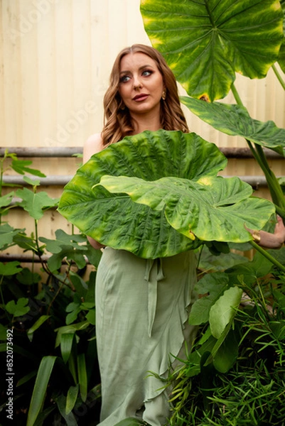 Obraz Cheerful blonde girl posing with sincere smile near big green leaf. Attractive fair-haired lady standing on nature background.