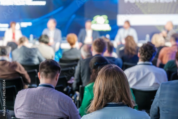 Fototapeta Business professionals in audience during a panel discussion at a conference. Shot from the back, focusing on engaged attendees.
