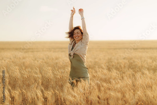 Fototapeta A happy, carefree woman with arms raised is celebrating in a vast golden wheat field on a sunny day, expressing joy and freedom.