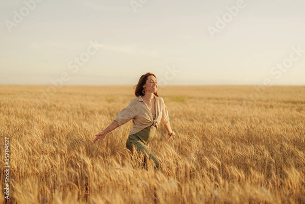 Fototapeta A happy young woman in casual attire enjoying a walk in a beautiful golden wheat field at sunset.
