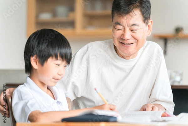 Obraz 家のダイニングで孫の宿題を教える高齢者男性と小学生の子供
