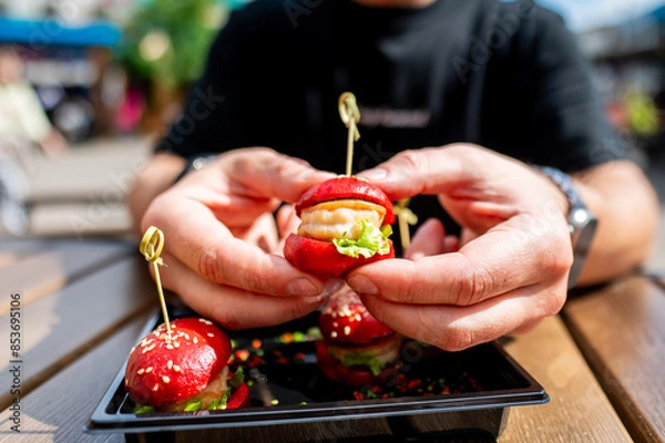 Fototapeta Close-up of hands holding a mini burger with colorful toppings, served on a black plate.