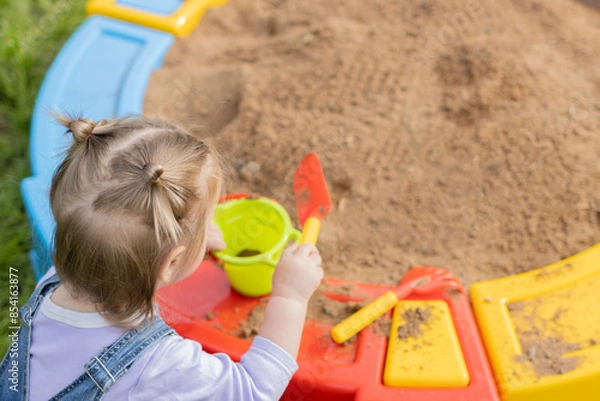 Fototapeta A young girl is playing in the sand box with a shovel and a cup.