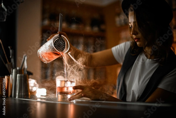 Fototapeta Woman bartender in a white t-shirt and vest pours a cocktail with ice from a mixing glass into another glass