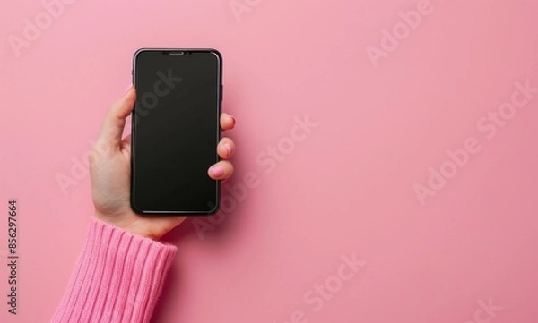 Fototapeta Young adult woman hand holding smartphone and touching black screen with finger on light pink table background. Pastel color. Empty place for text.