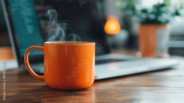 Fototapeta A steaming orange coffee mug on a wooden desk, with a blurred laptop and plant in the background, creating a cozy home office atmosphere.