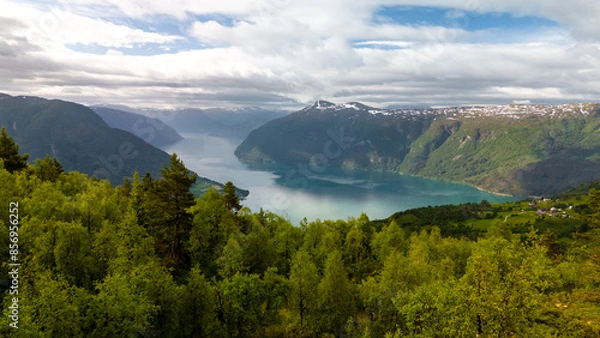Fototapeta A breathtaking view of a Norwegian fjord, surrounded by lush green forests and snow-capped mountains, under a cloudy sky. Molden hike, Norway