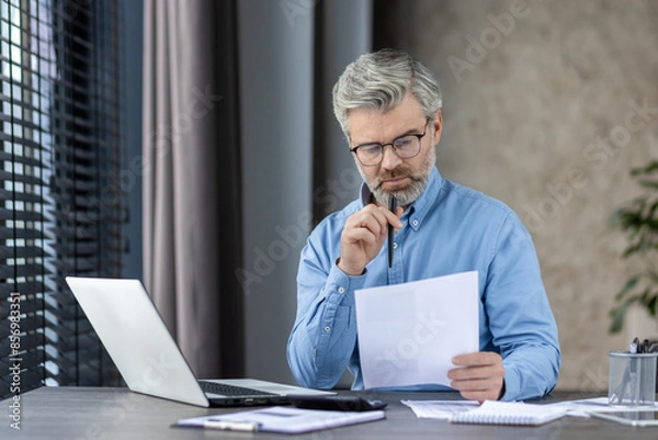 Fototapeta Senior businessman with gray hair and beard working from home or office, reading documents and using a laptop.