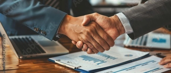 Fototapeta A pair of business partners shaking hands over a finalized financial plan, documents and charts visible
