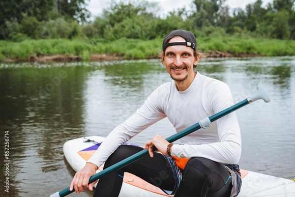 Fototapeta A man enjoys paddleboarding on a calm river, surrounded by nature. Outdoor water sports provide a serene and leisurely activity for an active lifestyle, making him happy