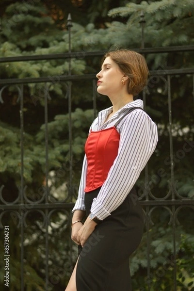 Fototapeta Stylish attractive woman in a white shirt and red top against a background of evergreen trees.