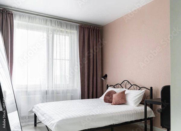 Obraz design of bedroom with beige walls, a big bed with a metal headboard, a white bedspread and pillows. There are flowers in a vase on the table in the bedroom, a chest of drawers and a mirror