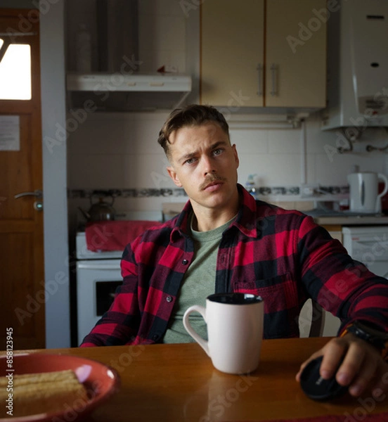 Obraz A young man with a mustache is sitting at a kitchen table. He has a cup of coffee in front of him and is looking at the camera.