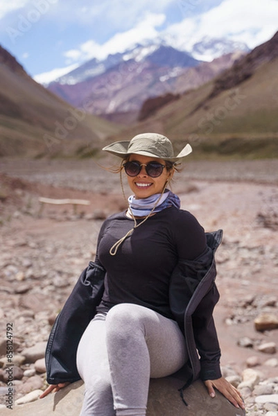 Obraz Portrait of a pretty young girl with a green mountain hat and sunglasses, sitting and smiling in front of the Aconcagua mountain.