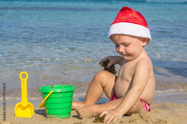 Fototapeta Baby by the sea in a cap of Santa Claus playing with sand