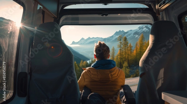 Fototapeta A person sits inside a van, admiring the golden-hour light over a mountainous landscape outside.