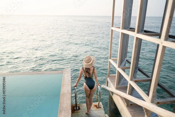 Fototapeta Woman in black swimsuit relaxing by infinity pool with ocean view.