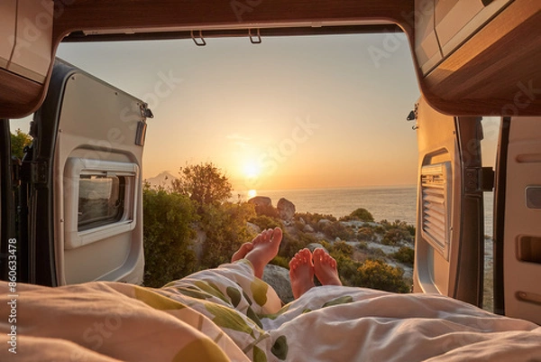 Fototapeta Couple enjoying a sunset view from their camper van bed, with toes emerging from under the covers, overlooking the sea. Perfect for travel, adventure, and romantic getaway themes.
