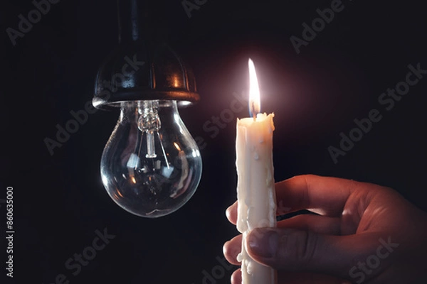 Fototapeta Burning candle near a switched off light bulb in complete darkness. Blackout, electricity off, energy crisis or power outage, concept image.