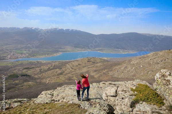 Fototapeta Mother and daughter at the top of the mountain pointing to the Pinilla reservoir, the Lozoya valley and the mountains of the Guadarrama National Park, in Madrid, Spain