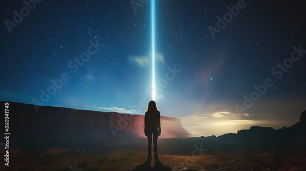 Fototapeta A woman stands in a desert at dusk, with a bright beam of light shooting straight up into the heavens behind her.