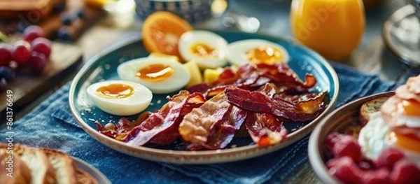 Fototapeta Copy space image of a breakfast spread featuring hard-boiled eggs, crispy bacon, fresh fruit, and toast on a blue placemat, with a horizontal layout and selective focus.
