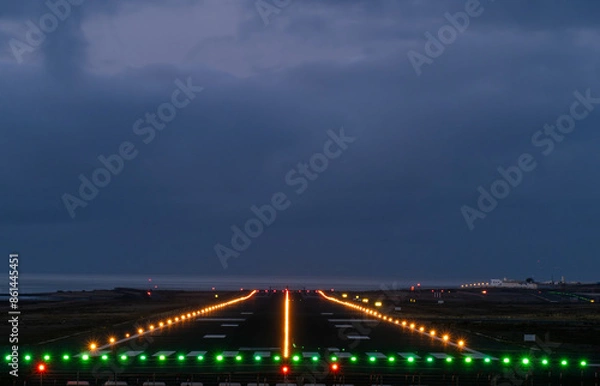 Fototapeta Runway illuminated with green and yellow lights at night with a view of the ocean in the background. The sky is dark and cloudy