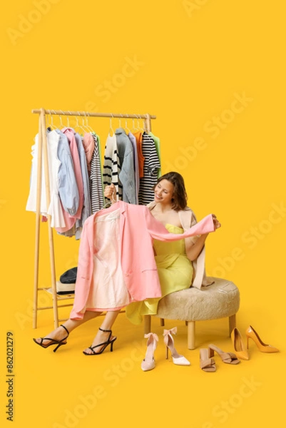 Fototapeta Beautiful young woman near rack with different clothes choosing blazer and shoes on yellow background