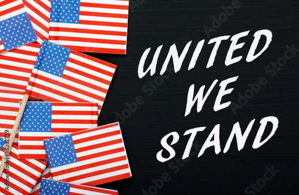 Fototapeta The phrase United We Stand in white text on a blackboard next to paper flags of the United States Of America