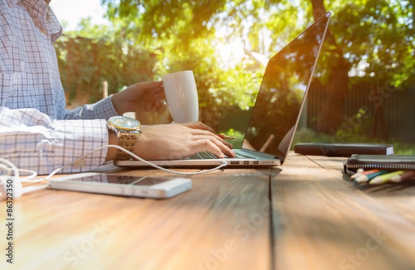Fototapeta Escaped of office.
Business style dressed man sitting at natural country style wooden desk with electronic gadgets around working on laptop drinking coffee sunlight and green terrace on background