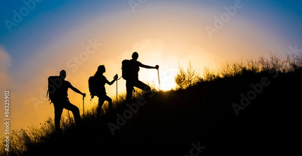 Fototapeta Wspinacze na trawiastym wzgórzu. Rodzina trzech osób sylwetka idąc strome trawiaste wzgórze majestatyczny wschód słońca i tło błękitnego nieba