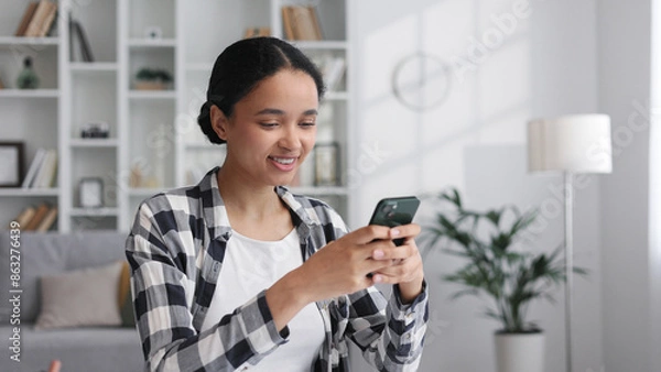 Obraz A young African American girl sits at the table in the living room, smiles, chats with friends on a mobile phone, happily taps the screen, reads happy news.