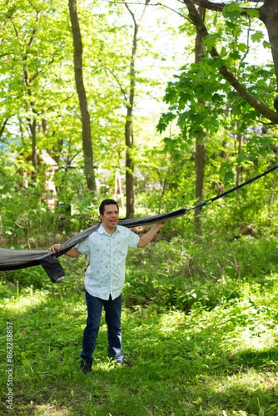 Fototapeta A young man holds a large black and gray hammock. The hammock is suspended between two trees. The man smiles and enjoys the fresh air.