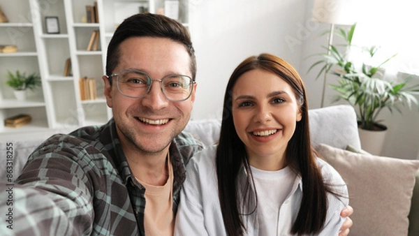 Obraz Happy young couple sitting on sofa in living room looking at camera conversation during video call at home, smiling, man and woman talking via webcam using unlimited internet connection.