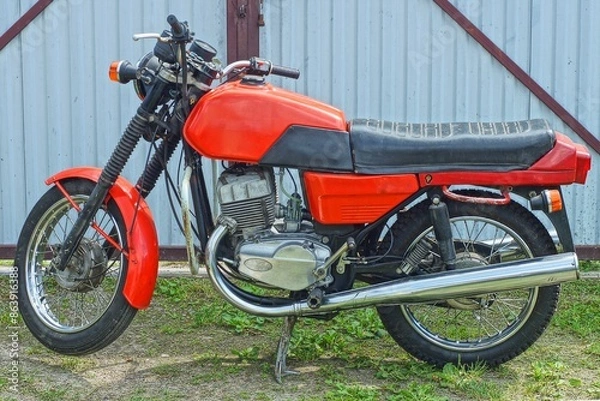 Fototapeta one red iron dirty old retro with two wheels industrial powerful heavy motorcycle stands on the ground near a gray fence during the day on the street