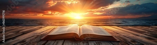 Obraz Open book on wooden surface with dramatic sunset background.