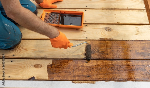 Fototapeta A worker wearing orange gloves carefully applies a dark brown stain to a wooden deck with a roller. The deck is partially stained, with the freshly stained wood gleaming in the sunlight.