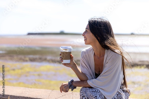 Fototapeta Young woman enjoying hot tea sitting in front of ocean on windy day 