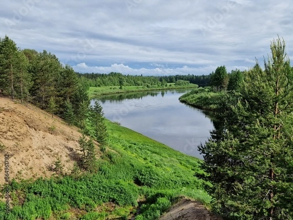 Fototapeta A Winding River Through A Lush Forest Landscape on a Cloudy Summer Day. The river is calm and reflective, reflecting the green trees and the cloudy sky. The forest is dense and green, with tall trees 
