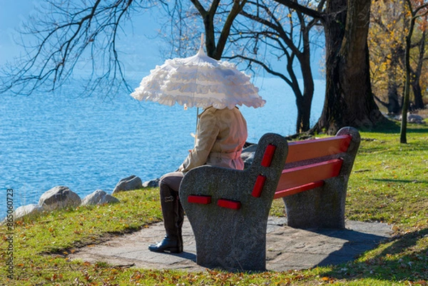 Fototapeta Woman with a Parasol Sitting on a Bench on the Waterfront to Lake Maggiore in a Sunny Day in Locarno, Ticino, Switzerland.