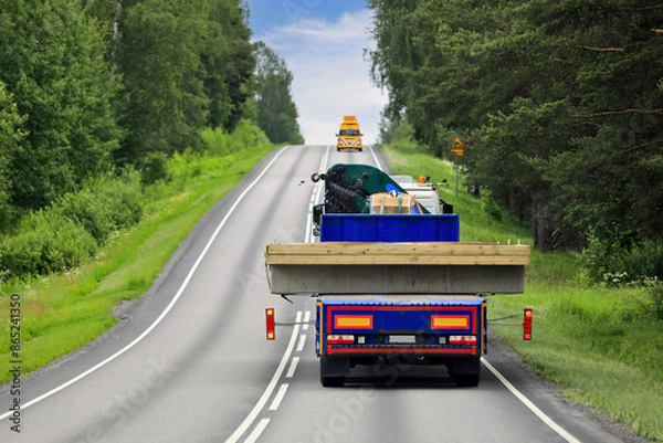 Obraz Heavy truck and trailer transporting wide load along highway with pilot vehicle ahead leading the way. Rear view, copy space.
