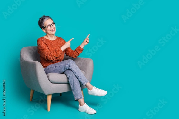 Fototapeta Full length photo of lovely pensioner lady sit armchair look point empty space dressed brown garment isolated on cyan color background