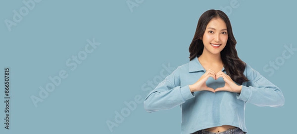 Fototapeta Smiling young asian woman showing heart gesture by using two hands and looking at camera isolated over blue background copy space 