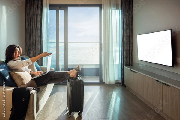 Obraz Mid aged asian businesswoman relax in home or hotel resting after travelling using laptop. Woman choosing movie on internet streaming service with remote control in hand. White screen LED TV isolated.