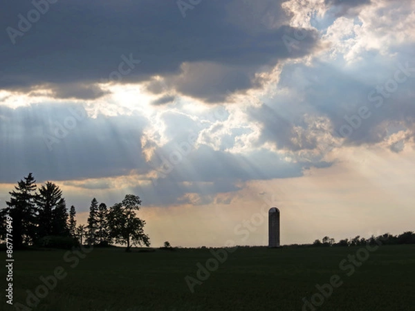 Obraz Trees and a lone silo are silhouetted by the sun beaming through the clouds. 