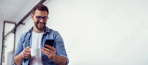 Obraz Handsome bearded man leaning on office desk, drinking coffee and talking on the phone.