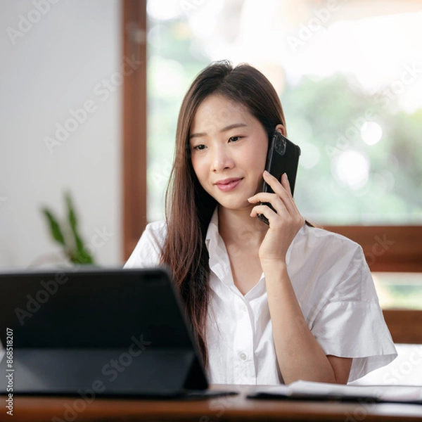 Obraz A woman is talking on her cell phone while sitting at a desk with a laptop