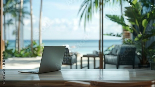 Fototapeta Tropical home office with ocean view. Laptop on a wooden desk with a blurred background of a tropical beach, perfect for travel and work-from-anywhere concepts.